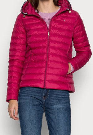 Nainen pukeutuneena kirkkaan pinkkiin, tikattuun vetoketjulliseen takkiin, jossa on huppu, päällään vaaleanvioletti neule ja siniset farkut.