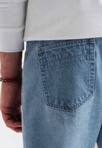 Primer plano de la mano de una persona que lleva una pulsera de cuentas junto al bolsillo trasero de unos pantalones vaqueros de mezclilla azul claro y una camisa blanca de manga larga.