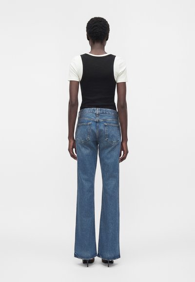 Άτομο που στέκεται γυρισμένο με την πλάτη, φοράει μαύρο αμάνικο τοπ πάνω από λευκό μπλουζάκι με κοντά μανίκια, μπλε τζιν με ψηλή μέση και φαρδιά μπατζάκια, και μαύρες ψηλοτάκουνες.