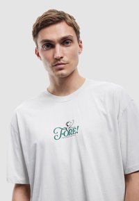 Camiseta de algodón blanca de manga corta, con un logotipo impreso en verde y un gráfico en el área del pecho izquierdo. Textura suave, ajuste relajado.
