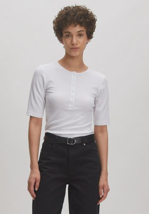 Weißes geripptes T-Shirt mit kurzen Ärmeln und Rundhalsausschnitt, ausgestattet mit einer Knopfleiste mit sechs Knöpfen. Kombiniert mit schwarzen Hosen und einem Gürtel.