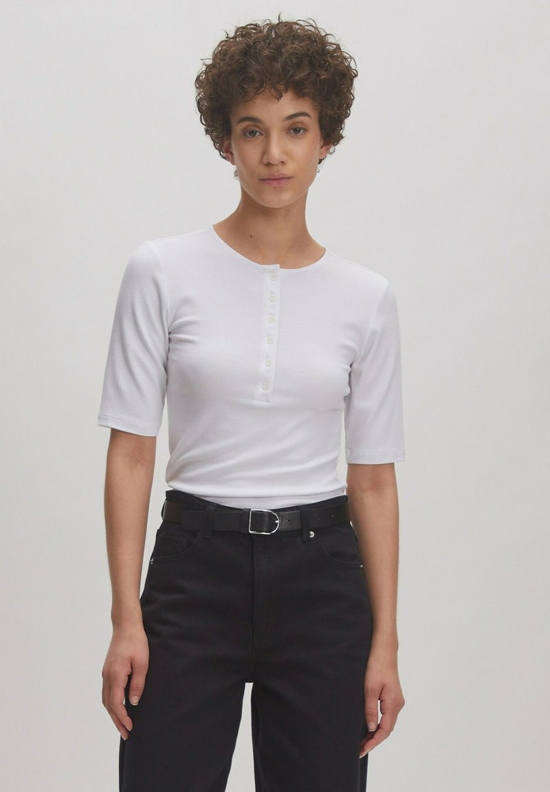 Witte geribde t-shirt met korte mouwen en een ronde hals, voorzien van een zesknopssluiting. Gepaard met zwarte broeken en een riem.