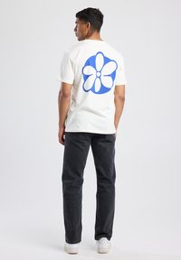 Crème t-shirt met een grote blauwe bloemenprint op de rug, gecombineerd met donkere jeans en witte sneakers, met een ontspannen pasvorm.
