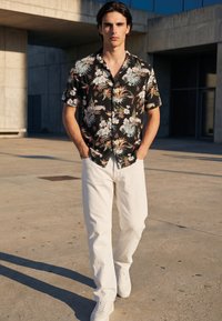 Ung mann som går ute, iført en svart blomstret kortermet skjorte, hvite bukser og hvite joggesko, hender i lommene.