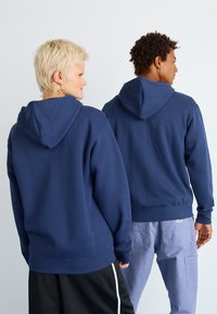 Sweatshirts azul-marinho em um tecido suave e texturizado, apresentando um capuz grande e punhos canelados, usados por duas pessoas vistas de costas.