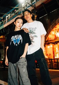Camiseta negra con gráfico de mariposa, combinada con pantalones cargo oliva y pantalones cargo grises. Camiseta blanca con una ilustración de un árbol.
