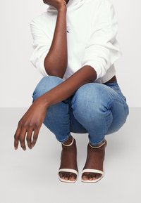 Vit huva sweatshirt, blå skinny jeans och ljus beige öppna klacksandaler. Har croppad design och exponerade vrister.