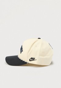 Béžová a černá baseballová čepice Nike s vyšitým nápisem "New York" na přední straně a logem Nike našitým na boku, položená na bílém povrchu.