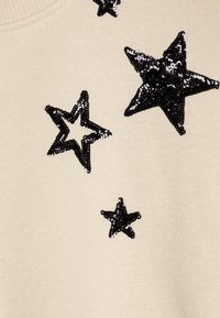 Jersey beis con adornos de estrellas de lentejuelas negras de diferentes tamaños, que presenta una textura suave y un cuello redondo.