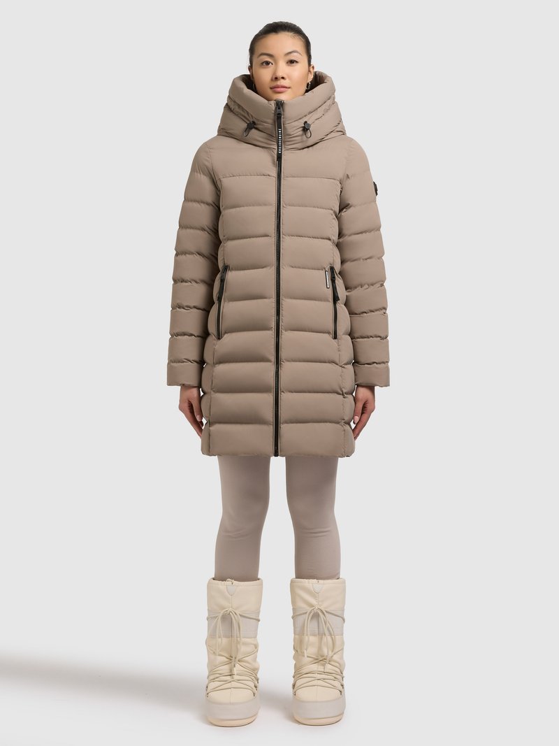 Beiger, wattierter Wintermantel mit hohem Kragen und Kapuze, ausgestattet mit einem Frontreißverschluss und zwei Seitentaschen. Kombiniert mit weißen Schneestiefeln und hellen Leggings.