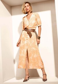 Maxikleid in hellem Orange mit Blumenmustern, kurzen Puffärmeln, Knopfleiste und einem braunen strukturierten Gürtel. Getragen mit braunen Absatzschuhen.
