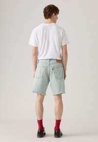 Shorts en denim bleu clair avec des ourlets effilochés, poches arrière et une étiquette en cuir marron. Portés avec un t-shirt blanc et des chaussettes rouges, associés à des chaussures noires.