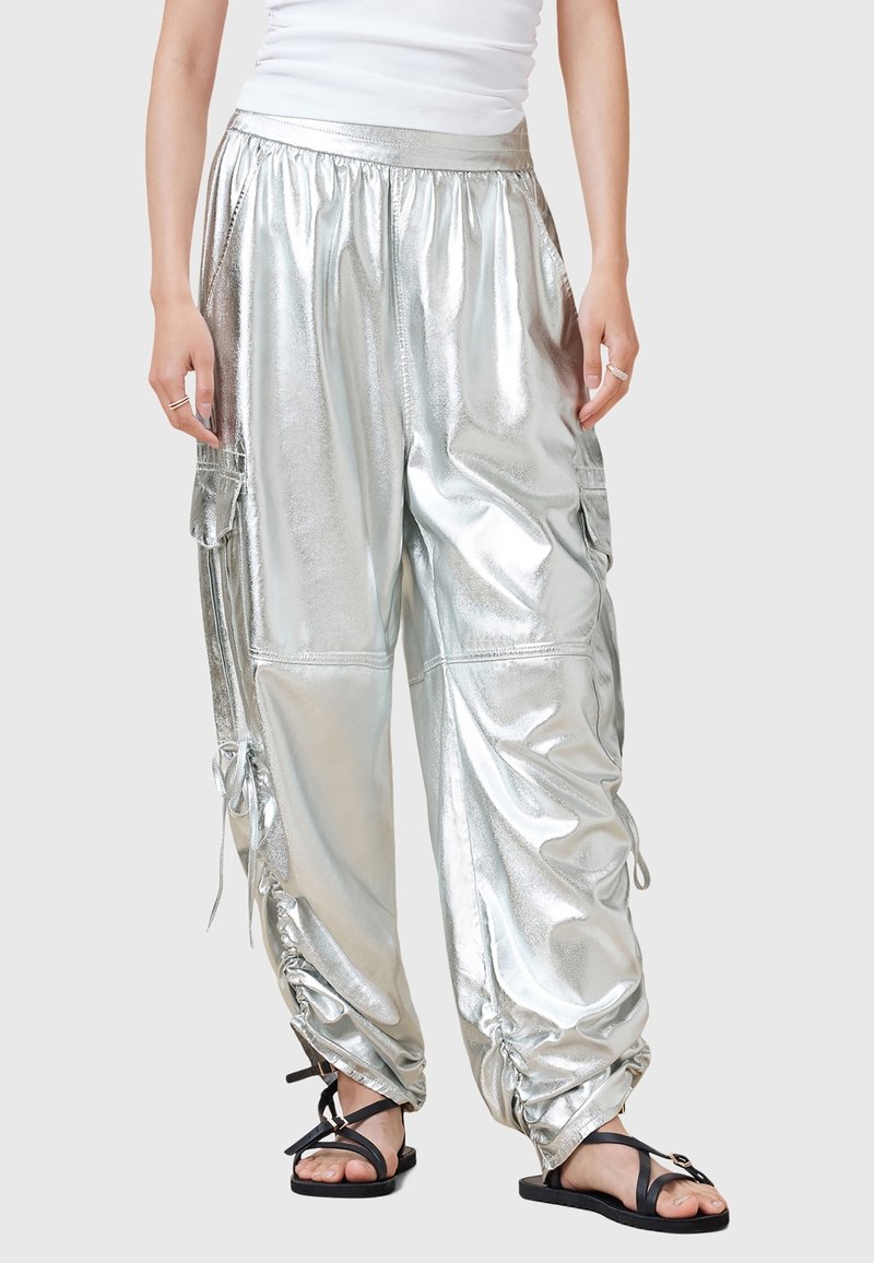 Personne portant un pantalon cargo argenté brillant et ample avec des cordons aux chevilles, associé à des sandales plates noires à brides et un haut blanc sans manches.