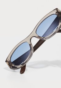 Óculos de sol oversized com uma armação cinza clara e lentes azuis. Apresenta um acabamento brilhante e hastes castanhas. O logótipo da marca é visível na lente.