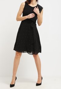 Musta hihaton mekko, jossa on vartalonmyötäinen yläosa, levenevä helma ja metalliset helmiäiskoristeet. Mukana sopiva vyötärönauha ja vuorattu kangas.