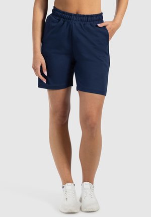 Donna che indossa pantaloni corti elasticizzati blu navy e sneakers bianche, in piedi con una mano in tasca contro uno sfondo semplice.
