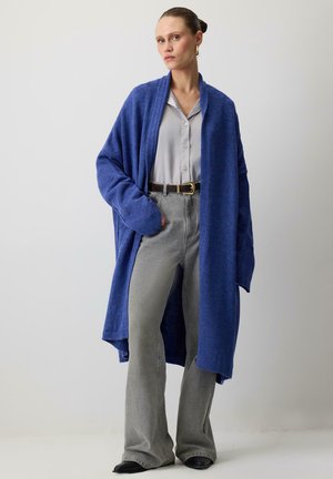 Kvinde iført en lang blå cardigan over en lys grå skjorte med knapper, grå vidde jeans, sorte sko, med håret samlet, stående opad en ensfarvet væg.