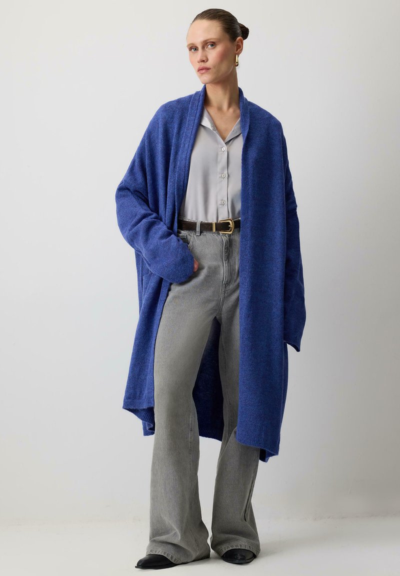 Naine, kellel on seljas pikk sinine kardigan heleda halli nööbivaba särgi, hallide laienevate teksade ja mustade kingadega, juuksed tagasi seotud, seismas lihtsa seina ees.