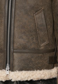 Hnedá kožená bunda s textúrovaným povrchom, vybavená strieborným zipsom a krémovým kožúškovým lemom pozdĺž spodného okraja.