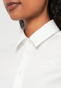 Balta krekla ar apkaklīti, izgatavota no gludas auduma, ar smalku izšūtu logotipu pa kreisi un pogu aizdari uz priekšējā slīdņa.