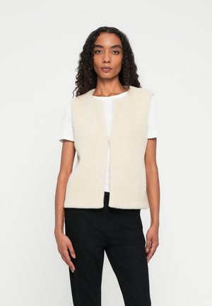 Fleece vest in cream color, featuring a round neckline, open front, and no sleeves. Paired with a white t-shirt and black pants.