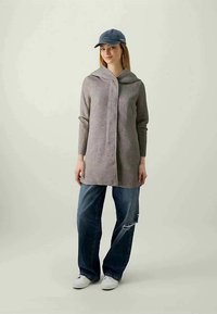 Camisola de manga longa cinza claro com capuz, fecho de botão e ajuste solto, combinada com jeans azul desgastado e tênis brancos.