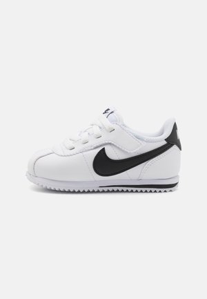 Hvid lav sneaker med sort Nike swoosh, snørelukning foran og tekstureret sål, vist fra siden på en hvid baggrund.