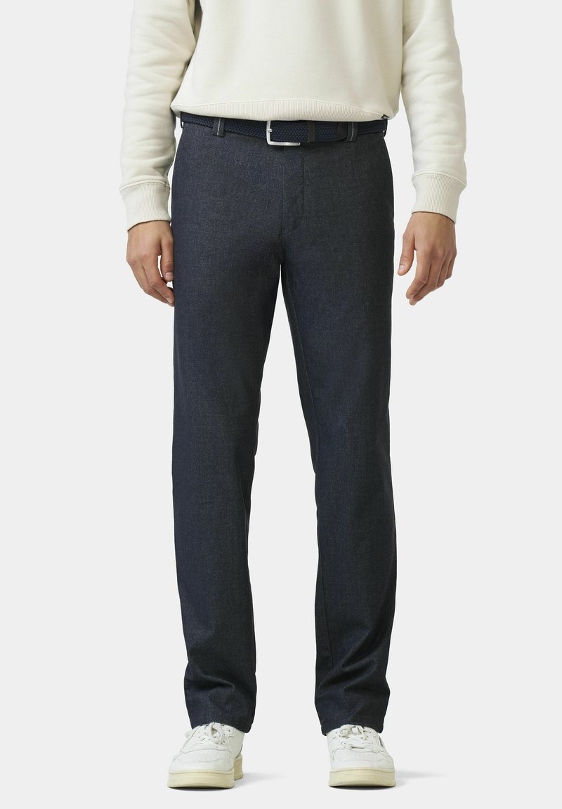 Persoon in donkerblauwe recht gesneden broek, witte sneakers, een gevlochten marineblauwe riem en een lichtbeige lange mouw top, staande tegen een effen achtergrond.