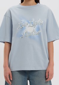 Svetle modré oversized tričko vyrobené z bavlny, s třpytivým nápisem "Low Lights Studios" na jemně gradientním pozadí. Krátké rukávy.