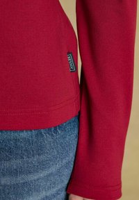 Nahaufnahme einer Person, die ein rotes Langarmoberteil mit einem "Joules"-Label und blauen Jeans trägt, vor einem neutralen Hintergrund.