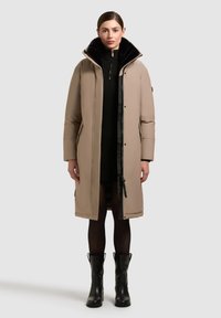 Beige párnázott parka fekete műszőrme gallérral, cipzáras elülső résszel, zsebekkel és térdig érő fazonnal, fekete kötött ruhára és fekete csizmára húzva.