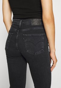 Vue arrière d'une personne portant un jean noir taille haute et un haut noir sans manches, avec des poches arrière visibles et une étiquette en cuir de la marque sur la ceinture.