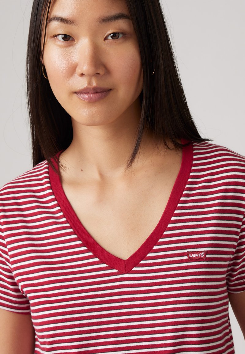 Camiseta de rayas rojas y blancas con cuello en V y mangas cortas. Presenta un pequeño parche del logo de Levi's en el pecho. Material de algodón, textura suave.