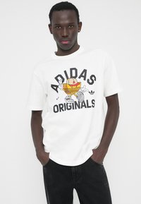 Baltas medvilninis marškinėlis su spalvotu grafiniu atspaudu su užrašu „ADIDAS ORIGINALS“, derinamas su tamsiomis džinsinėmis kelnėmis.