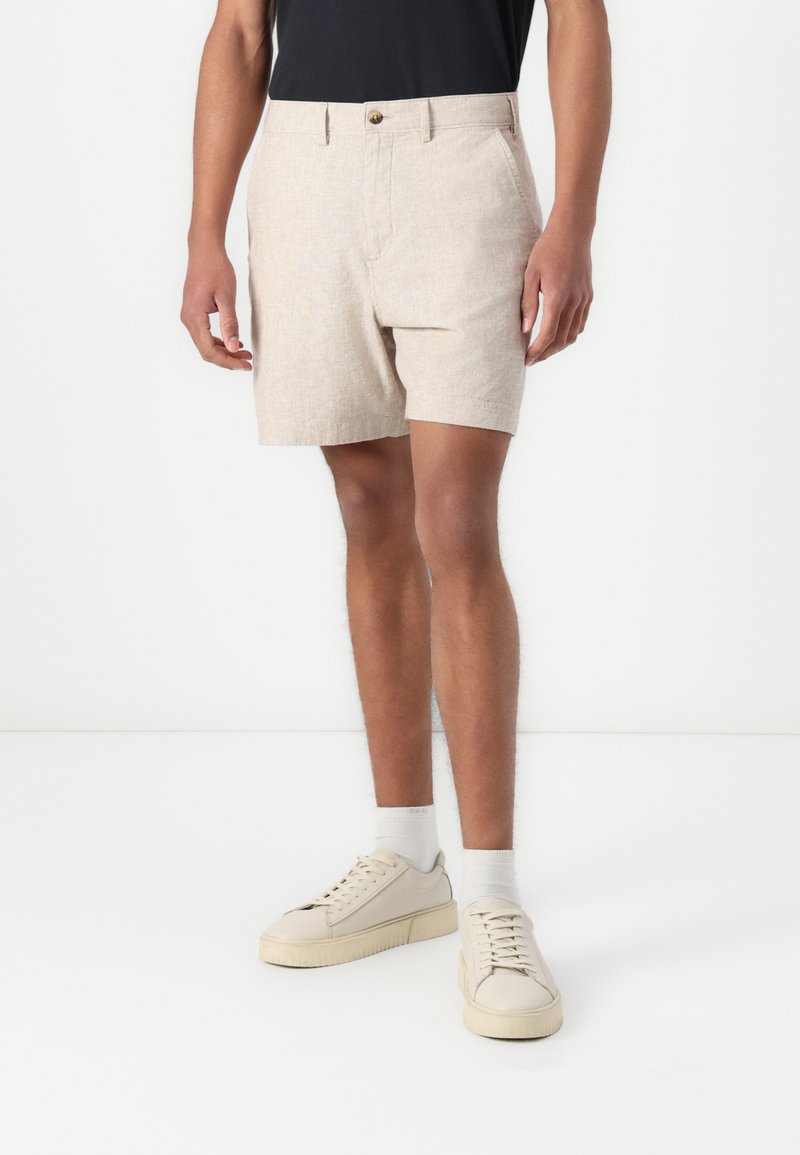 Shorts en lin beige avec une texture lisse, dotés d'une fermeture à bouton à l'avant et de poches latérales, associés à des baskets blanches.