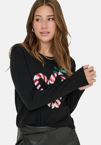 Schwarzer Pullover mit langen Ärmeln, der ein rotes und weißes Herzdesign in Form eines Zuckerstangenmusters zeigt, verziert mit Glitzer und einem grünen Schleifenakzent.