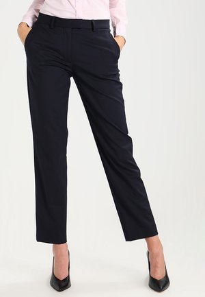 Vrouw die zwarte op maat gemaakte broek met zijzakken en zwarte hoge hakken draagt, handen in de zakken, en een lichtroze overhemd.