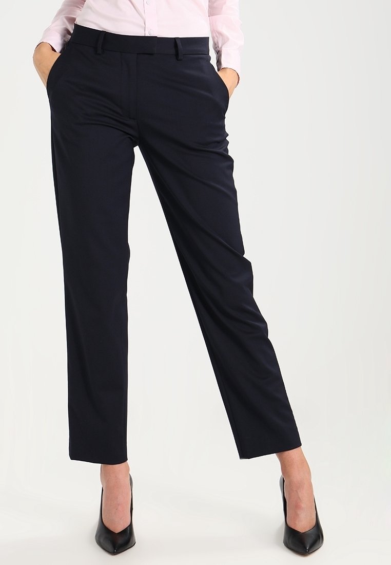 Femme portant un pantalon noir sur mesure avec poches latérales et des chaussures à talons noirs, mains dans les poches, et une chemise rose clair boutonnée.