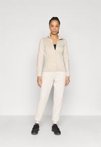 Veste beige à fermeture éclair avec col montant, associée à un pantalon de survêtement beige clair. Chaussures de sport noires complètent la tenue. Texture de tissu lisse.