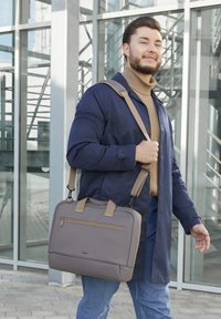 Mann in einer blauen Jacke und hellbraunem Rollkragenpullover geht vor einem Glasm gebä ude und trägt eine graue Umhängetasche.