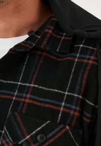 Camicia a quadri nera con linee rosse, blu e bianche. Realizzata in lana, presenta un colletto, dettagli con bottoni e una tasca sul petto.