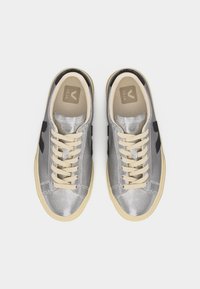 Pár stříbrných nízkých tenisek s krémovými tkaničkami, béžovými podrážkami a černým logem "V" na vnitřních stranách, pohled shora na bílém pozadí.