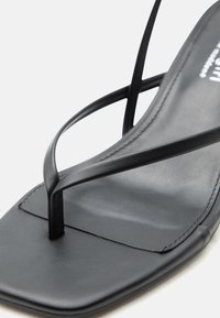 Μαύρο δερμάτινο σανδάλι με λεπτά σταυρωτά λουράκια και σχεδιασμό με τετράγωνη μύτη, εμφανιζόμενο σε κοντινό πλάνο με λευκό φόντο.