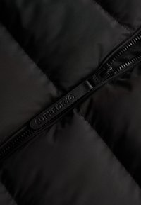 Svart pufferjacka i tyg med quiltad textur. Har en kontrasterande dragkedja och en upphöjd, präglad logotagg på dragkedjan.