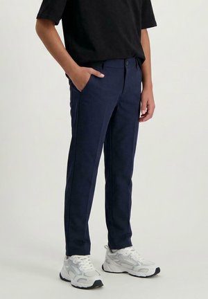 Persoon draagt marineblauwe slim-fit broek en witte sportsneakers, staat met één hand in de zak tegen een effen achtergrond.