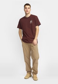 Bordeaux katoenen T-shirt met korte mouwen, voorzien van een klein wit grafisch ontwerp. Gecombineerd met beige cargobroeken en beige schoenen.