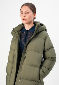 Vrouw in een olijfgroene gewatteerde jas met capuchon over een marineblauw shirt met kant en kraag, die iets opzij kijkt.