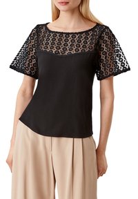 Haut noir avec un corps lisse et des manches en dentelle noire présentant un motif floral. L'encolure est arrondie, avec un design légèrement transparent.