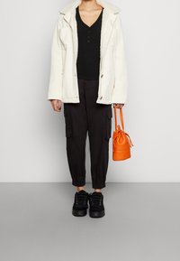 Giacca bianca con collo in shearling, maglia nera, pantaloni cargo neri, sneakers nere con plateau e borsa arancione piccola.