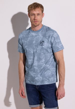 Blonder Mann mit leichtem Bart, der ein blau gemustertes T-Shirt mit Palmenlogo und hochgekrempelte dunkle Jeansshorts trägt, steht vor einer weißen Wand.